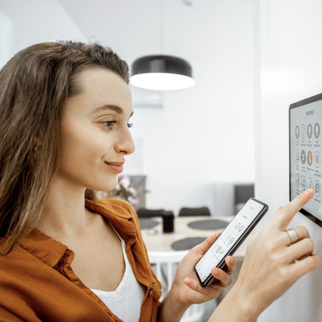 Woman using smart home interface on a wall-mounted screen while holding a smartphone.