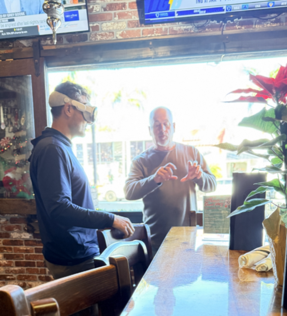 A person wearing a mixed-reality headset stands at a bar while another person gestures with their hands, appearing to explain an interface or interaction; daylight streams through a window behind them.