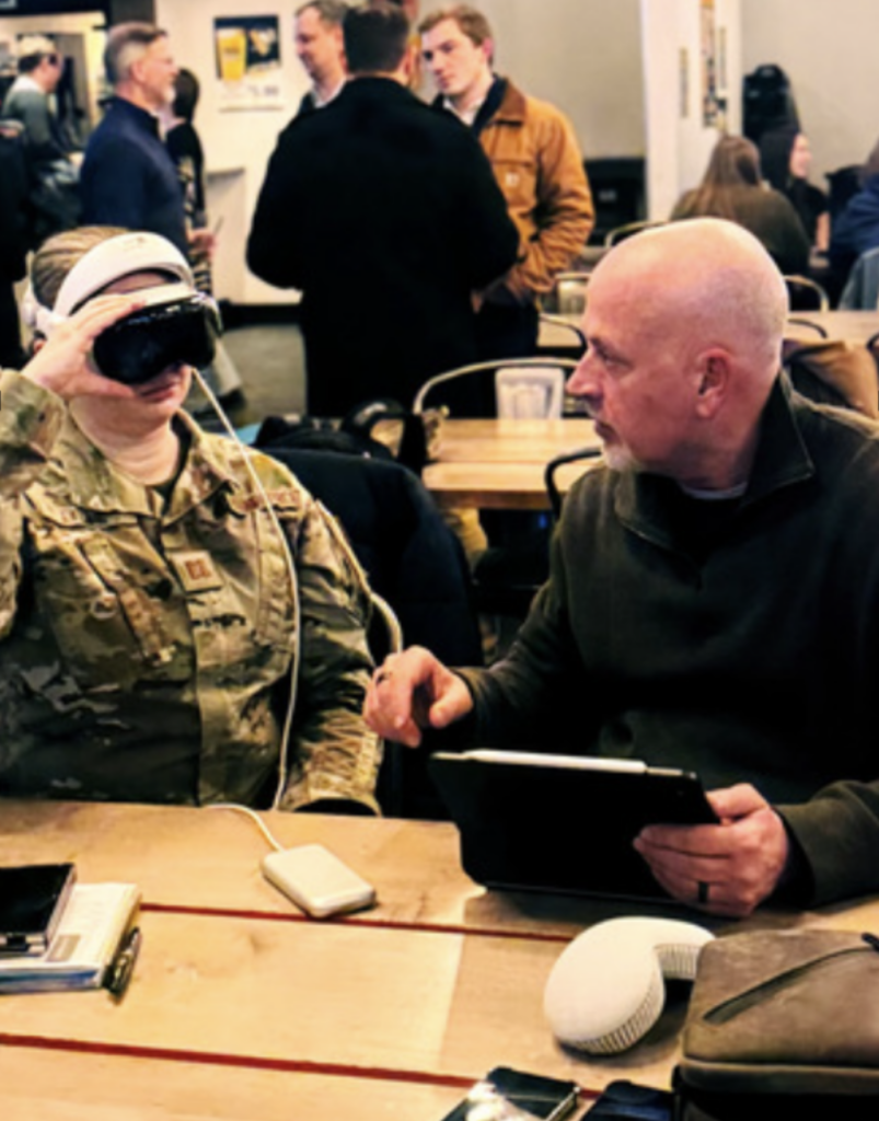 A person in military camouflage wears a VR headset while seated at a table, as another person beside them gestures and explains something; several people talk in the background.